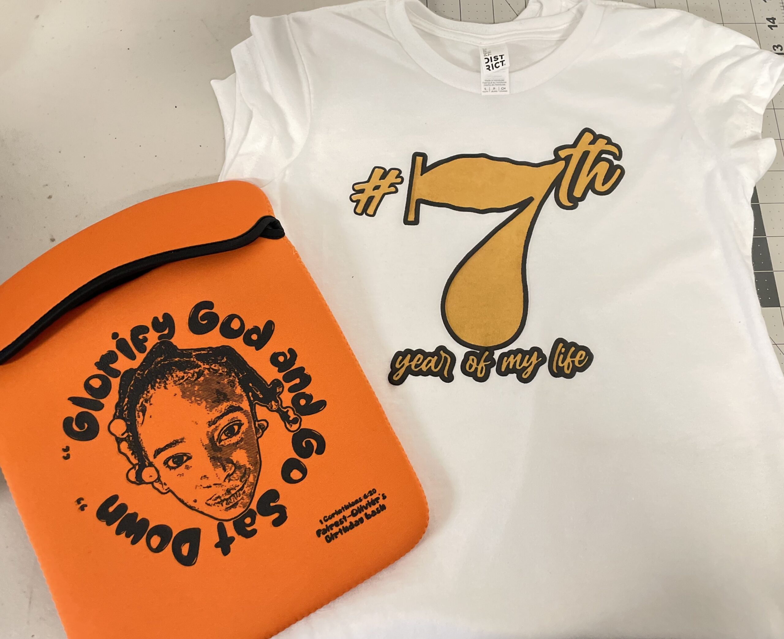 A white tee with a large gold “7th” graphic is paired with a bright orange printed accessory featuring a portrait and circular lettering.