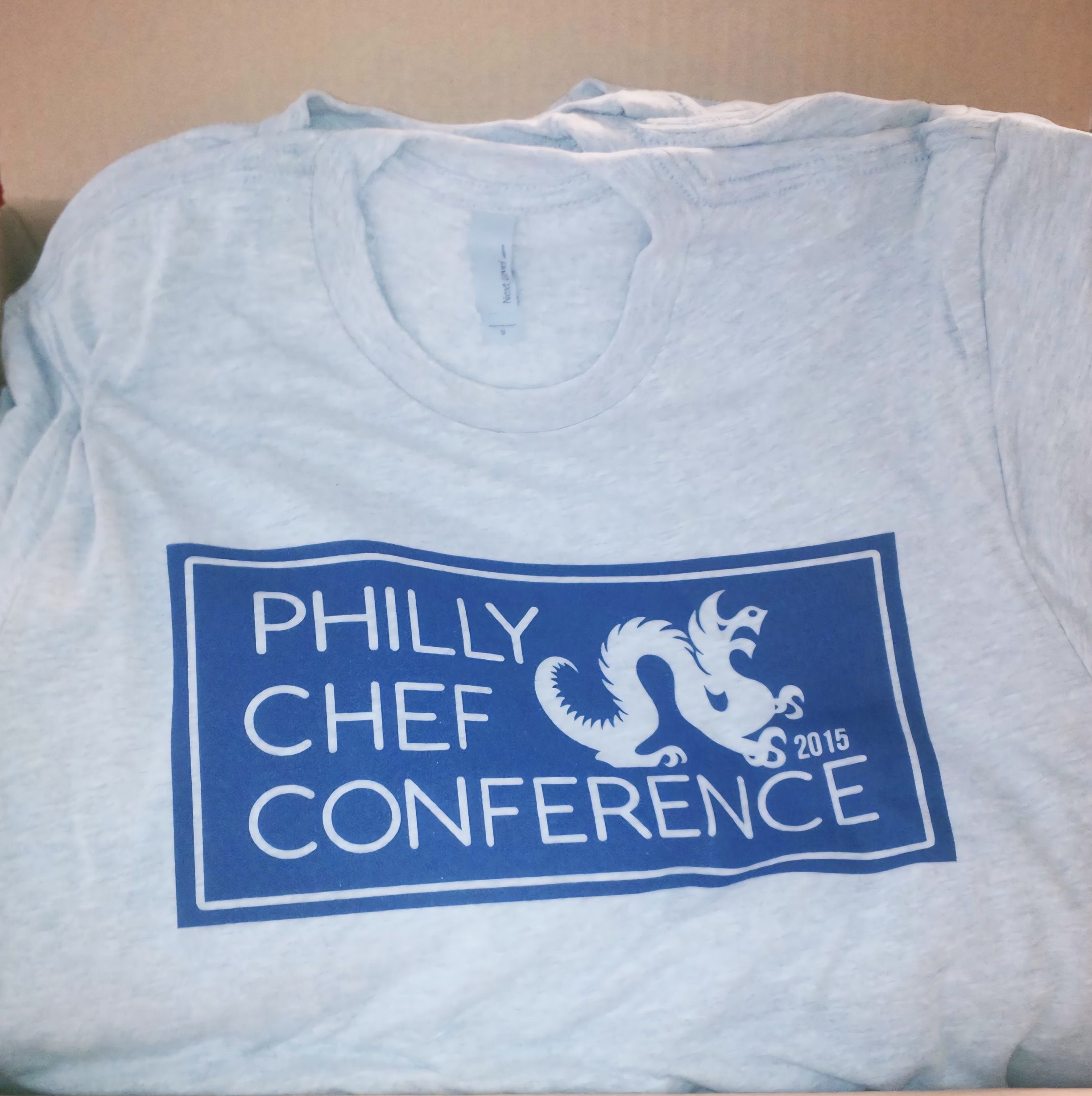 A pale blue shirt displays white Philly Chef Conference lettering inside a blue rectangular print, photographed flat to show the design.