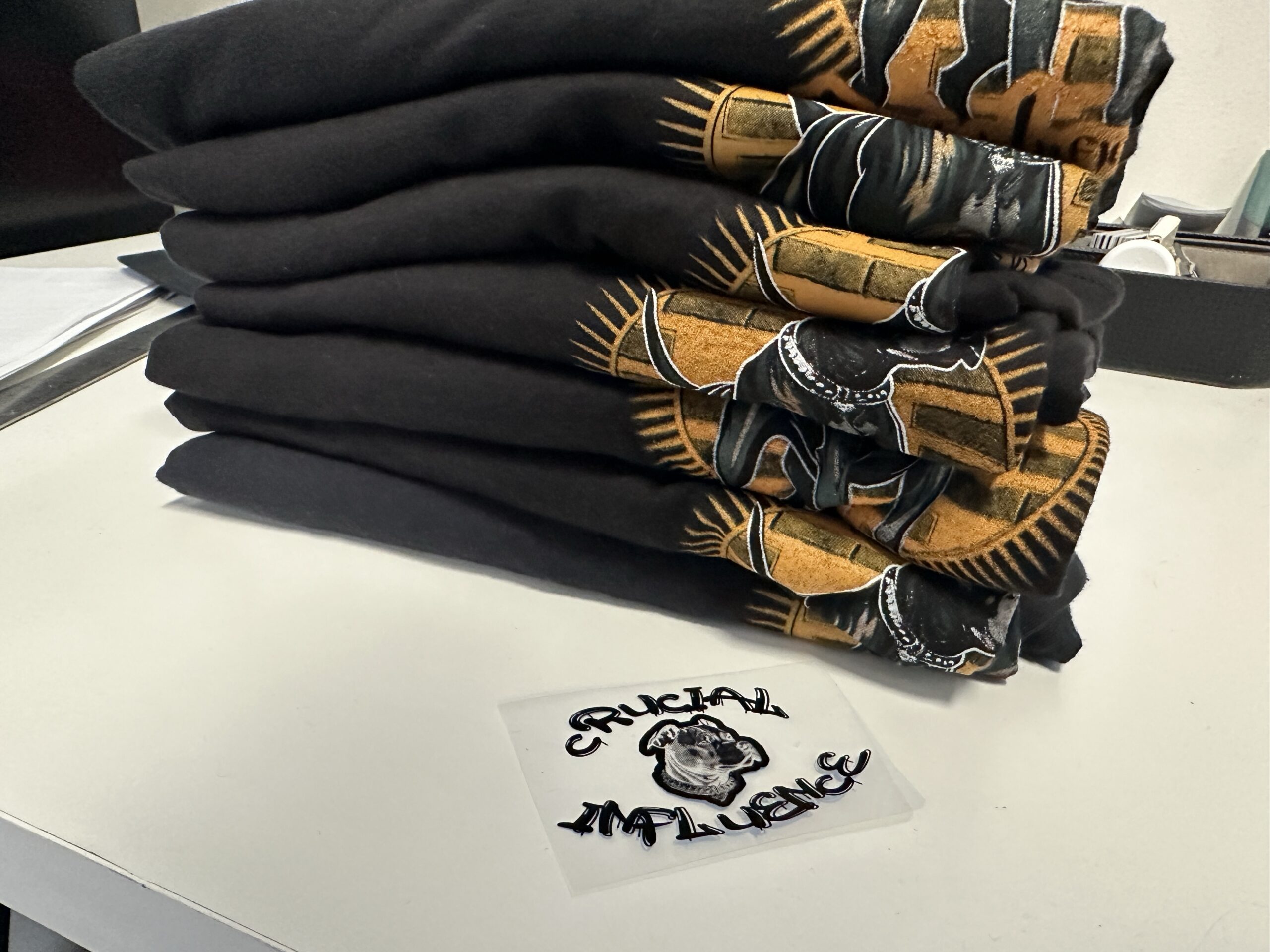 A stack of dark printed garments sits above a light-colored piece. Repeated gold artwork shows several finished prints together.