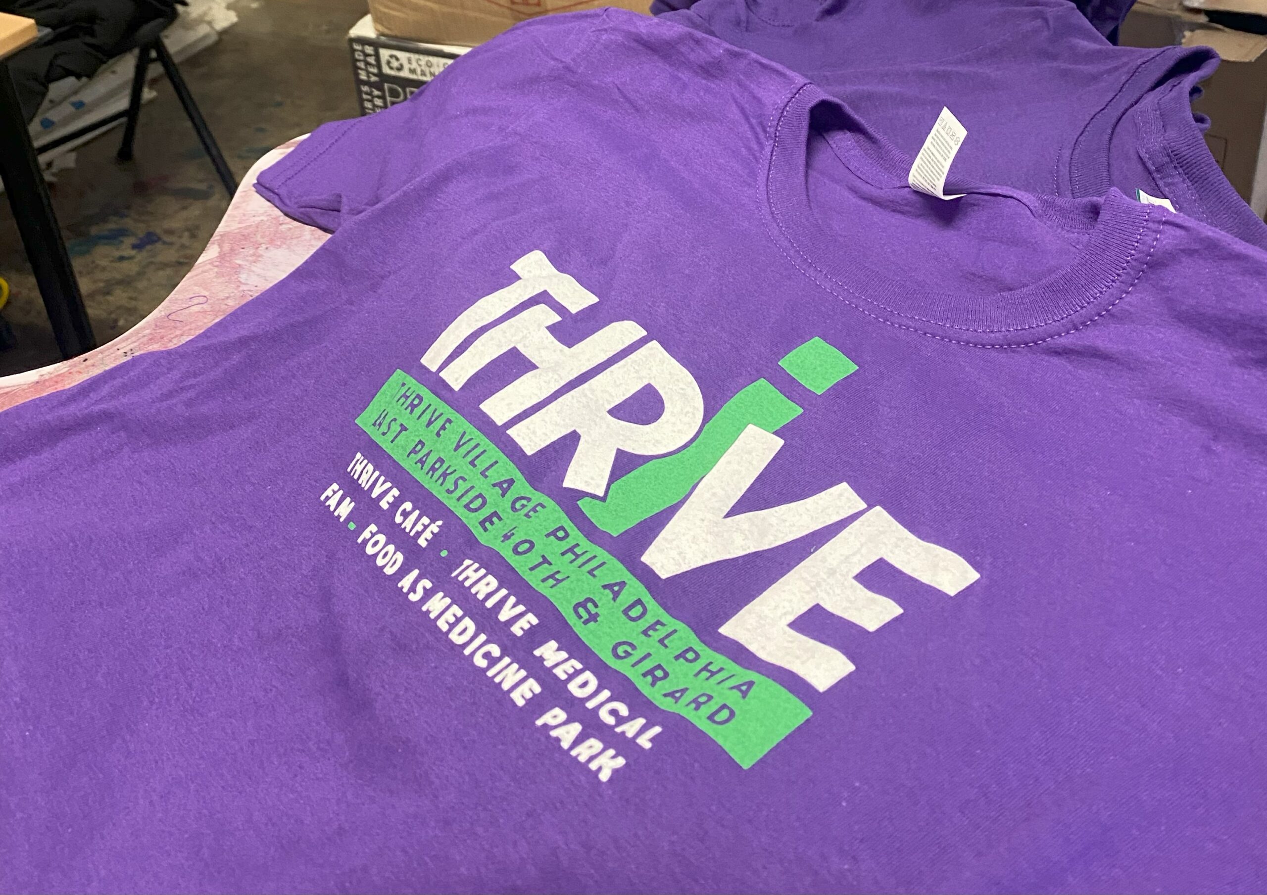 A purple shirt features large white “THRIVE” lettering, a green accent and smaller event text below.